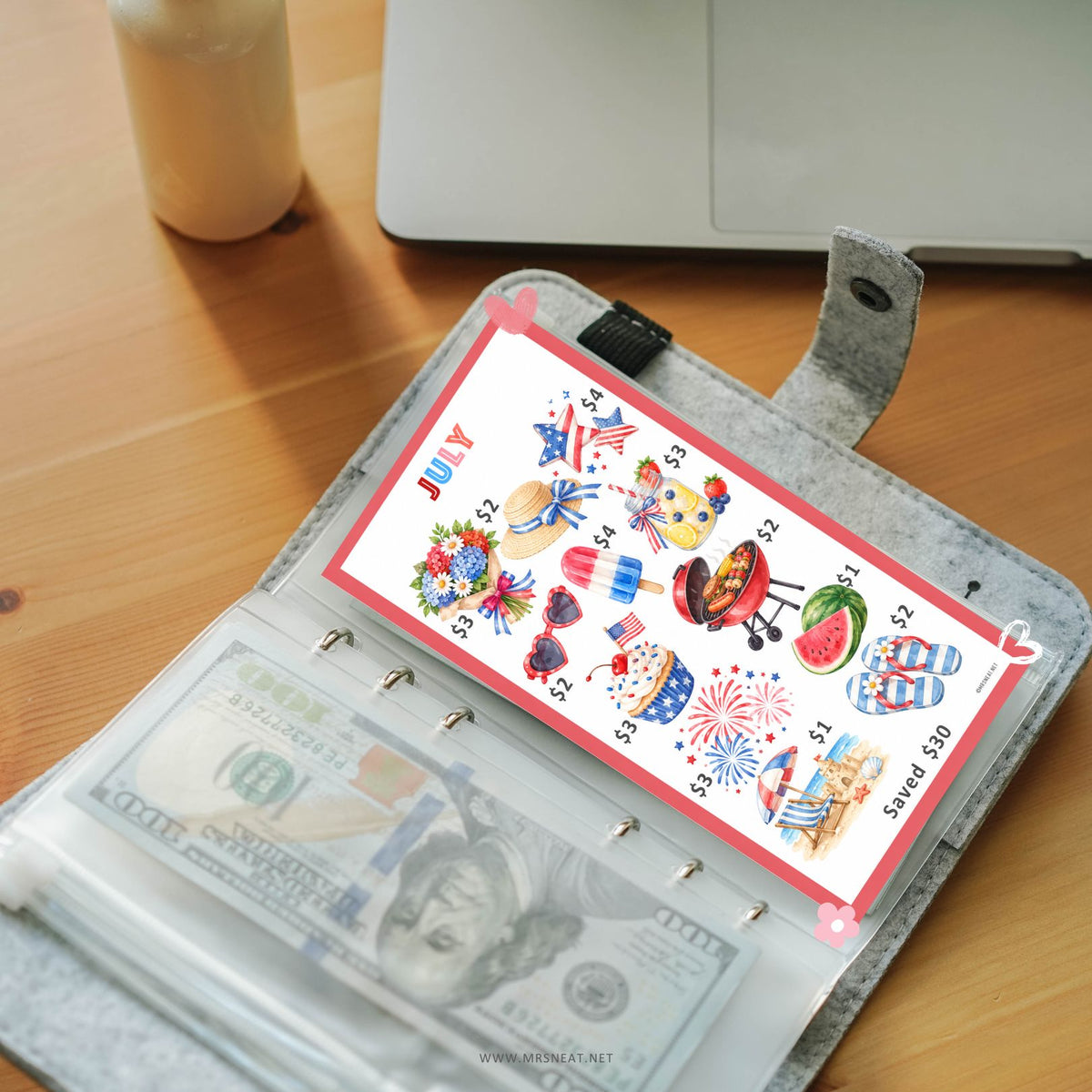 A ring binder planner or wallet in a gray felt-like material, open on a wooden desk. Inside is a white sticker sheet with patriotic summer-themed illustrations such as fireworks, a grill, watermelon, sandals, and the word 'JULY,' plus cash in a clear sleeve.